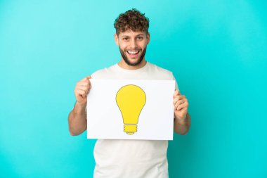 Mavi arka planda izole edilmiş yakışıklı beyaz bir adam elinde mutlu ifadeli bir ampul simgesi olan bir levha tutuyor.