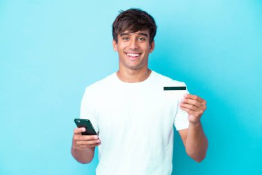 Kafkasyalı genç adam mavi arka planda izole edilmiş cep telefonuyla kredi kartıyla alışveriş yapıyor.