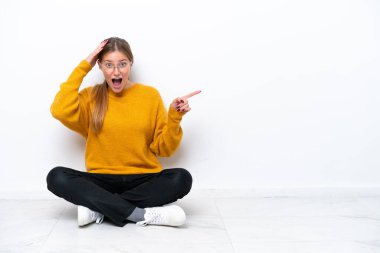 Beyaz arka planda izole bir şekilde yerde oturan genç beyaz kadın şaşırdı ve parmağını yana çevirdi.