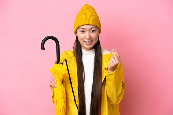 Young Chinese woman with rainproof coat and umbrella isolated on pink background making money gesture