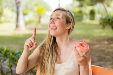 Açık havada donut tutan genç sarışın kadın parmağını kaldırarak çözümü anlamaya çalışıyor.