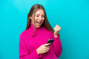 Genç Litvanyalı kadın mavi arka planda izole edilmiş ve telefon zafer pozisyonunda.