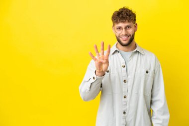 Sarı arka planda izole edilmiş yakışıklı beyaz adam mutlu ve parmaklarıyla dört kişi sayıyor.