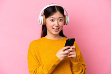 Genç Çinli bir kadın pembe arka planda izole edilmiş. Cep telefonuyla müzik dinliyor ve ön tarafa bakıyor.