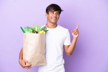 Mor arka planda izole edilmiş bir alışveriş çantası tutan genç adam harika bir fikre işaret ediyor.