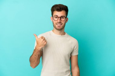 Mavi arka planda izole edilmiş beyaz, yakışıklı genç bir adam bir ürün sunmak için kenarı işaret ediyor.