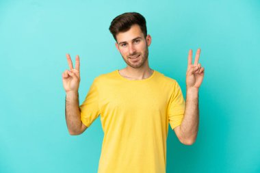 Mavi arka planda izole edilmiş beyaz, yakışıklı genç bir adam iki eliyle zafer işareti gösteriyor.