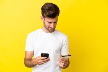 Genç beyaz yakışıklı adam sarı arka planda izole edilmiş cep telefonuyla kredi kartıyla alışveriş yapıyor.
