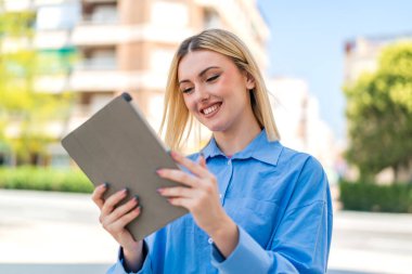 Açık havada güzel sarışın bir kadın elinde mutlu bir ifadeyle bir tablet tutuyor.