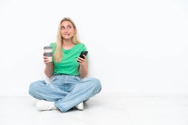 Beyaz arka planda izole bir şekilde yerde oturan beyaz tenli genç bir kadın elinde kahve ve bir şeyler düşünürken bir cep telefonu tutuyordu.