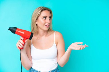 Mavi arka planda saç kurutma makinesi tutan beyaz bir kadın. Şaşırtıcı bir yüz ifadesi var.