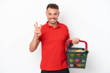 Beyaz arka planda izole edilmiş yiyecek dolu bir sepet taşıyan beyaz bir adam mutlu ve parmakları olan üç kişi sayıyor.