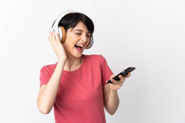 Kısa saçlı bir kadın beyaz arka planda izole edilmiş. Cep telefonuyla müzik dinliyor ve şarkı söylüyor.