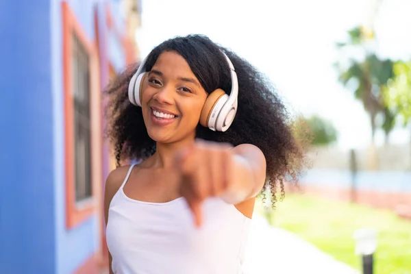 Young African American woman with headphones at outdoors points finger at you with a confident expression