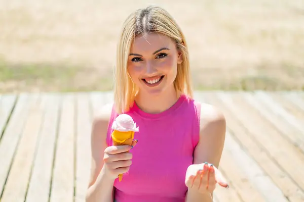 Young pretty blonde woman with a cornet ice cream at outdoors inviting to come with hand. Happy that you came