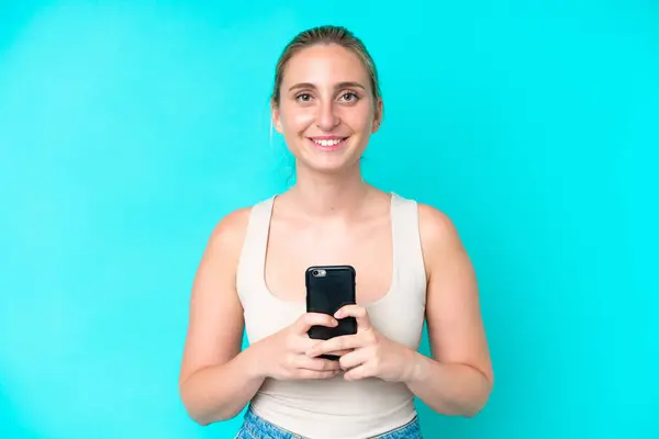 携帯電話を使いながらカメラを見て微笑む青い背景に孤立した若い白人女性 — ストック写真