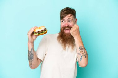 Kızıl saçlı, uzun sakallı, elinde burgerle mavi arka planda zaferi kutlayan bir adam.