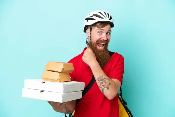 Delivery man holding pizzas and burgers isolated on blue background celebrating a victory