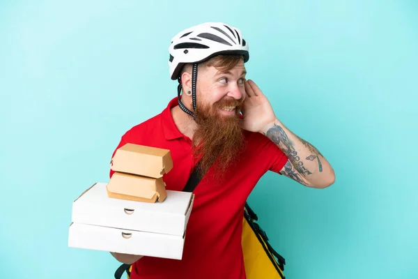 Delivery man holding pizzas and burgers isolated on blue background listening to something by putting hand on the ear
