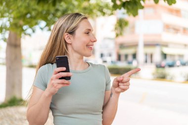 Açık havada cep telefonu kullanan genç sarışın bir kadın bir ürün sunmak için yan tarafı işaret ediyor.