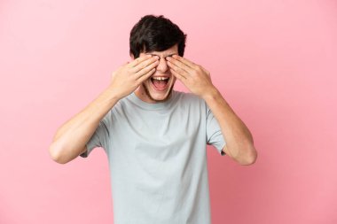 Pembe arka planda izole edilmiş genç bir Rus. Gözleri elleriyle kaplıyor ve gülümsüyor.