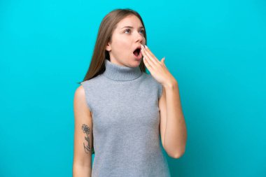 Genç Litvanyalı bir kadın mavi arka planda izole edilmiş esniyor ve ağzını eliyle açık tutuyor.