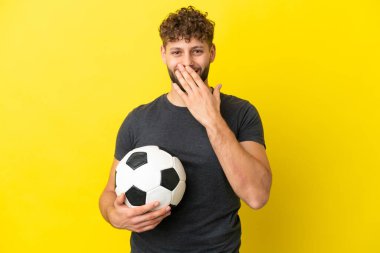 Yakışıklı genç futbolcu sarı arka planda izole edilmiş mutlu ve gülümseyen elleri ile ağzını kapatıyor.
