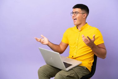 Genç adam dizüstü bilgisayarlı bir sandalyede oturuyor ve yüzünde sürpriz bir ifade var.