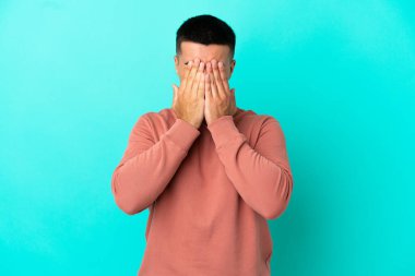 İzole edilmiş mavi arka planda genç yakışıklı adam gözlerini elleriyle kapatıyor.