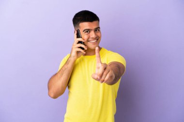 İzole edilmiş mor arka plan üzerinde cep telefonu kullanan genç yakışıklı bir adam parmağını kaldırıyor ve gösteriyor.