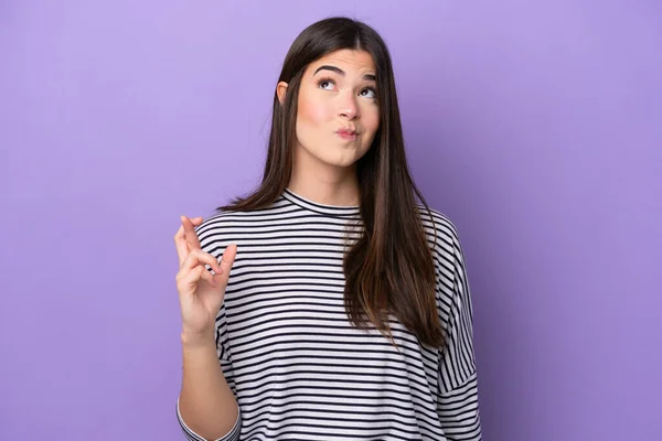 Young Brazilian woman isolated on purple background with fingers crossing and wishing the best