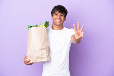 Elinde alışveriş çantası tutan genç adam mor arka planda izole edilmiş mutlu ve parmaklarıyla üçe kadar sayıyor.