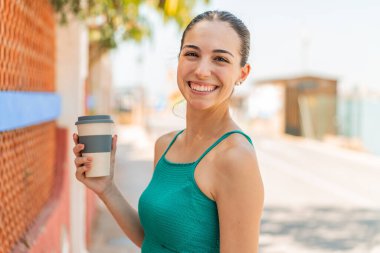 Genç ve güzel bir kadın elinde kahveyle dışarı çıkıp gülümsüyor.