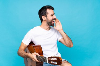 İzole edilmiş mavi arka planda gitar çalan genç bir adam ağzı sonuna kadar açık bağırıyor.