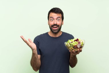Şok yüz ifadesi ile izole yeşil duvar üzerinde salata ile genç yakışıklı adam