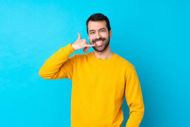 İzole edilmiş mavi arka planda genç beyaz bir adam telefon jesti yapıyor. Bana geri dönüş işareti ver.