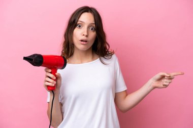 Pembe arka planda saç kurutma makinesi tutan beyaz bir kadın şaşırmış ve işaret ediyor.