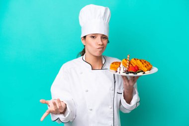 Kafkasyalı genç aşçı kadın mavi arka planda izole edilmiş waffle tutuyor. Omuzlarını kaldırırken şüpheyle hareket ediyor.