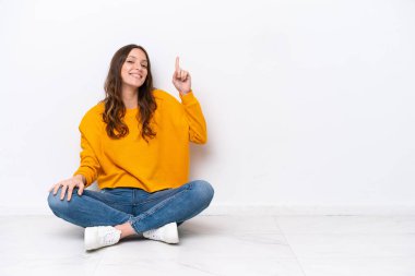 Beyaz duvarda izole bir şekilde yerde oturan genç beyaz kadın en iyinin işareti olarak parmağını kaldırıyor.