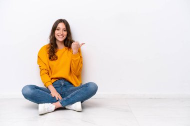 Beyaz duvarda izole bir şekilde yerde oturan genç beyaz kadın bir ürünü sunmak için yan tarafı işaret ediyor.