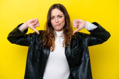 Sarı arka planda izole edilmiş genç beyaz kadın baş parmağını iki eliyle aşağı gösteriyor.