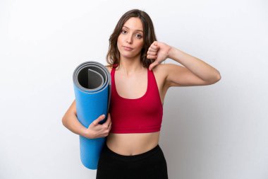 Beyaz arka planda izole edilmiş bir minderi tutarak yoga derslerine giden genç beyaz kadın negatif ifadeli baş parmağını gösteriyor.