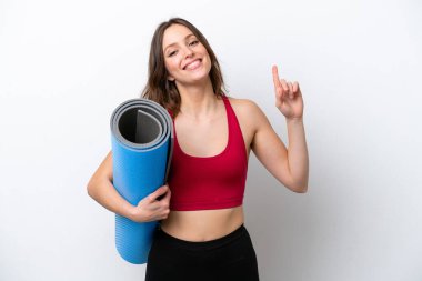 Beyaz arka planda izole edilmiş bir minderi tutarken yoga derslerine giden beyaz tenli genç bir kadın en iyinin işareti olarak parmağını kaldırıyor.