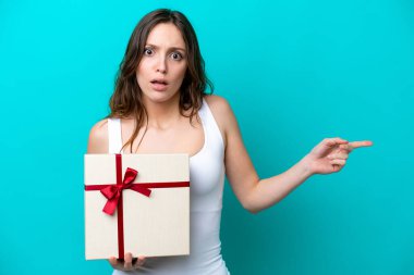 Young caucasian woman holding a gift isolated on blue background surprised and pointing side