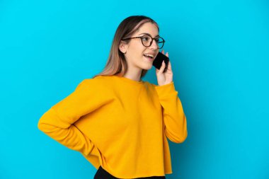 Mavi arka planda izole edilmiş genç beyaz kadın cep telefonuyla sohbet ediyor.
