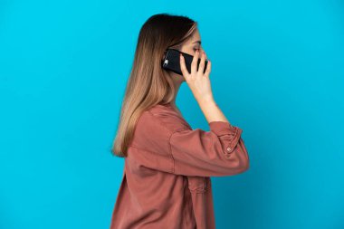 Mavi arka planda izole edilmiş genç beyaz bir kadın cep telefonuyla sohbet ediyor.
