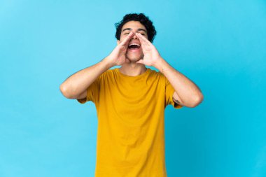 Genç Venezuelalı bir adam mavi arka planda izole edilmiş bağırıyor ve bir şey duyuruyor.