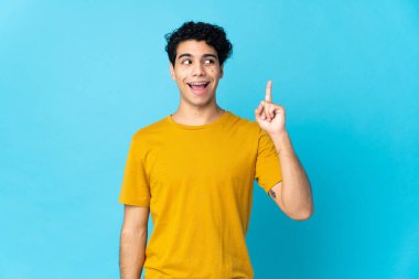 Genç Venezuelalı bir adam mavi arka planda izole edilmiş bir şekilde parmağını kaldırarak çözümü bulmaya çalışıyor.