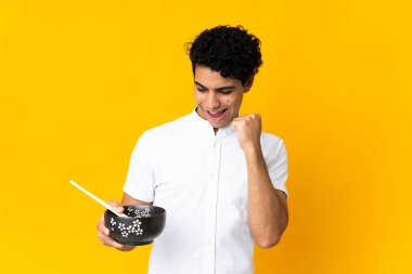 Genç Venezuelalı bir adam sarı arka planda izole edilmiş bir şekilde zaferini kutlarken elinde yemek çubuklarıyla bir kase erişte tutuyordu.
