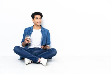Venezuelalı bir adam kahve almak için yerde oturuyor ve bir şeyler düşünürken cep telefonunu da yanında taşıyor.